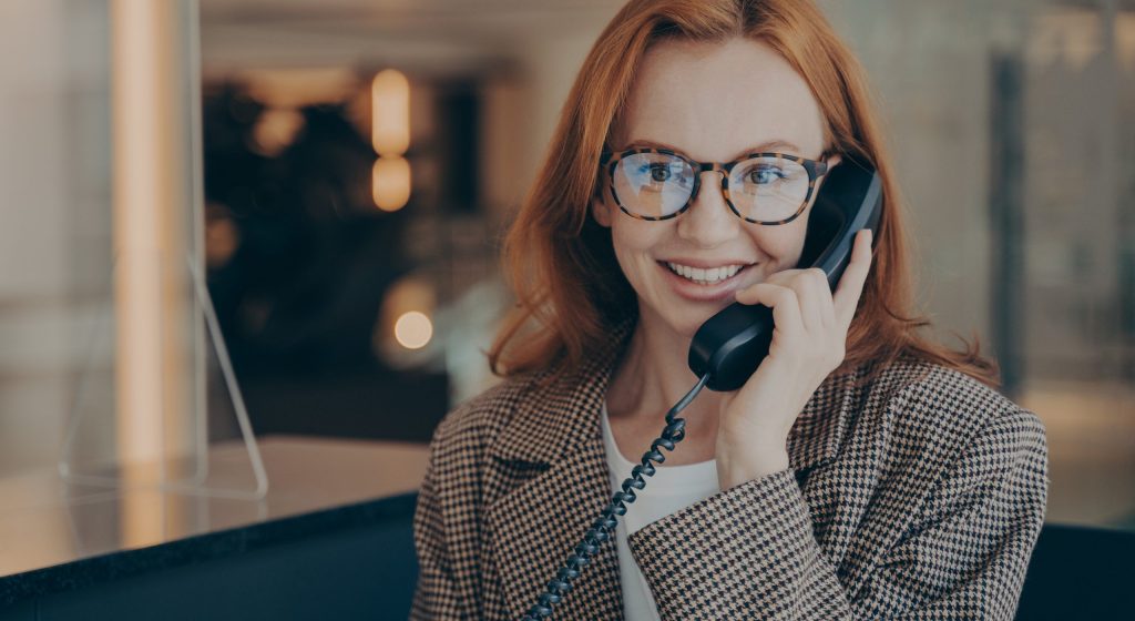 Smiling female office worker with red hair in plaid jacket wearing glasses, talking on landline phone with her co-worker about project completion date, office environment in blurred background